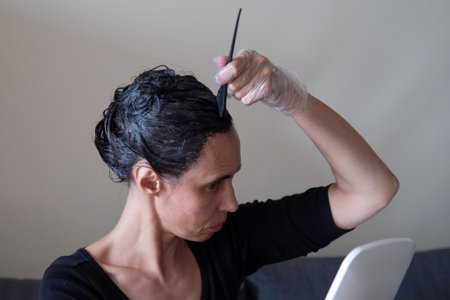 Middle age woman colouring dark hair with gray roots at home. Woman sitting on couch and dyeing hair...