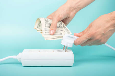 Woman hands holding electric power plug and US dollar banknotes near power strip extension cord on...