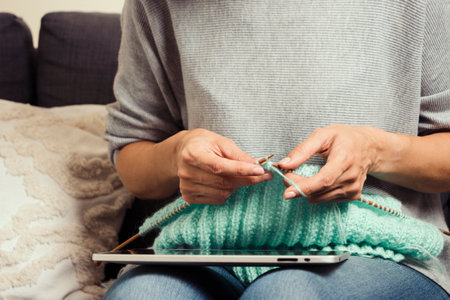 Middle age woman at home look at digital tablet computer screen and knitting sweater. Woman learning...
