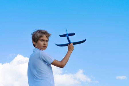 Child throwing toy plane. Kid holding plane and looking direct to the camera. Boy playing with blue toy airplane against blue sky on sunny day outdoorsの写真素材