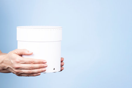 Woman hands holding white round cardboard box with cap on light blue background. Mock up package box. Front viewの写真素材