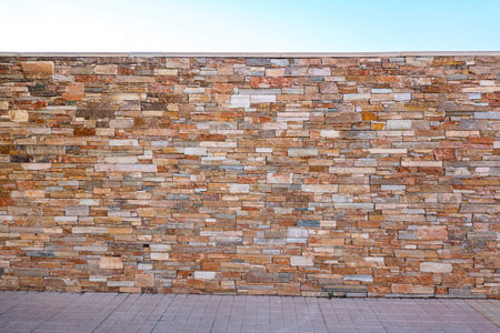Brown masonry fence of stones, pavement and blue sky. Light brown brick wall background with...