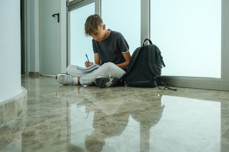 Teenage boy student wearing casual clothes sitting on floor and writing, makes notes in notebook....