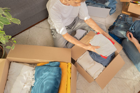 Mother and son sorting clothes and packing into cardboard box. Donations for charity, help low income families, declutter home, sell online, moving moving into new home, recycling, sustainable livingの写真素材