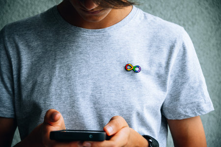 Teenage boy with autism infinity rainbow symbol sign metallic pin brooch on t-shirt using smartphone. World autism awareness day, autism rights movement, neurodiversity, autistic acceptance movementの写真素材
