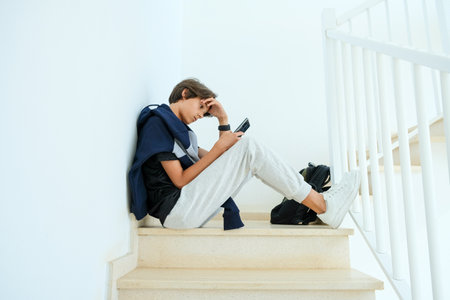 Teenage boy using smartphone. Confused, frustrated teenage boy sitting alone on stairs. Learning difficulties, gaming addiction, emotion, psychological problems in adolescence conceptの写真素材
