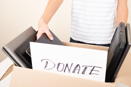 Donate technology to charity. Old used laptop computers, phone, tablet near cardboard box. Volunteer hands holding paper with text Donation. Donation box for e-waste, old tech equipment, reuseの写真素材
