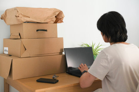 Woman working on laptop at home surrounded by parcel boxes. Small business owner preparing packages...