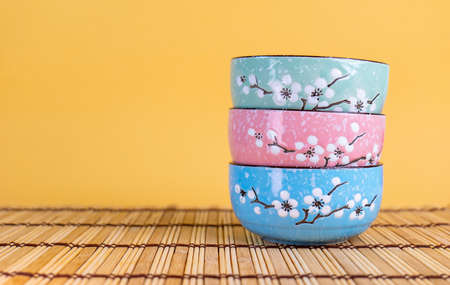 Three colored ceramic rice cups stacked in green, pink, blue on a placemat in a yellow scene.