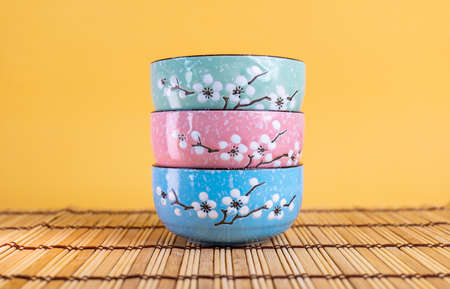 Three colored ceramic rice cups stacked in green, pink, blue on a placemat in a yellow scene.の写真素材