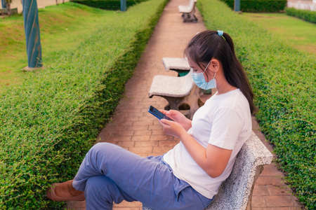 An Asian woman reading long-recorded messages on her mobile phone reminds me of the past. In the park wearing a mask to protect it for safety.の写真素材