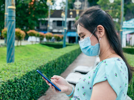An Asian woman reading long-recorded messages on her mobile phone reminds me of the past. In the park wearing a mask to protect it for safety.の写真素材