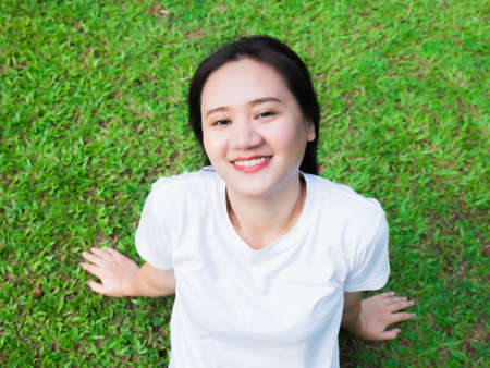 Asian girl in white shirt Come out to relax and enjoy the warm sunlight in the evening On a happy rest day from work Outside sitting on the green lawn, looking up at the light and smiling in the parkの写真素材