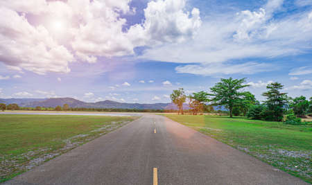 The road leads directly to the mountain view with lawns and trees on both sides. There is a wind blowing, cloudy sky and sunlight. There is a light flare. Ghost is a beautiful view and background.の写真素材