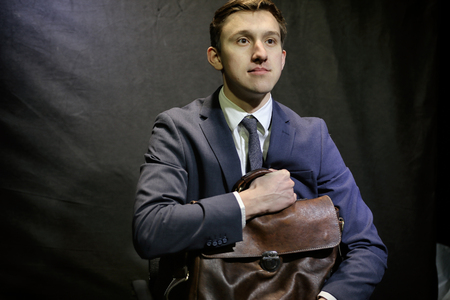 Young man in dark blue business suit sits with suitcaseの写真素材