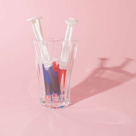 Red and blue syringe in glass on pink background. Minimal and pandemic concept.