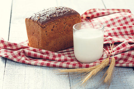 Freshly baked bread with spikelets of wheat and glass of milk on a red and white cloth on a wooden boards. close upの写真素材