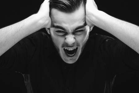 Angry young man, wearing in black shirt, is holding his hands on his head and screaming, on dark studio background, close upの写真素材