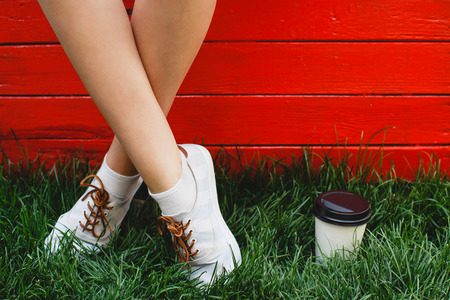 Feet of young girl, in white socks and sneakers, standing on green grass near the cup of coffee, on red wooden background, close upの写真素材