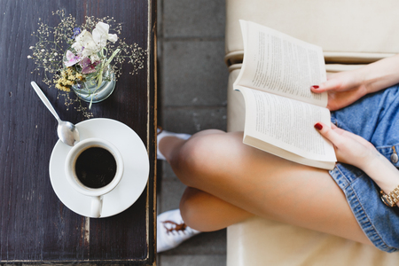 Young woman, wearing on blue shorts, sitting on beige leather sofa and holding a book, cup of coffee and vase with flowers on the dark wooden table, on cafe, close up. Point of viewの写真素材