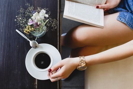 Young girl, wearing on blue shorts, sitting on beige leather sofa, reading a book and holding a cup of coffee, vase with flowers  standing on the dark wooden table, on cafe, close up. Point of viewの写真素材