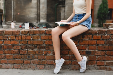 Slim young girl, wearing on white shirt, blue shirts and sneakers, sitting on a brick wall and reading a book, with cup of coffee and sunglasses on background, on the street, close up. Point of viewの写真素材