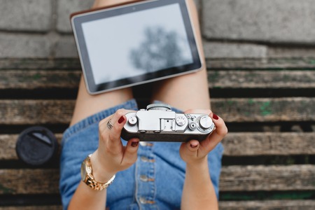 Young girl, wearing in blue shorts, sitting on a dark wooden bench with cup of coffee and tablet, and holding a camera,  close up. Point of viewの写真素材