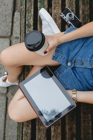 Slim young woman, wearing in blue skirt and sneakers, sitting on a wooden bench with cup of coffee...