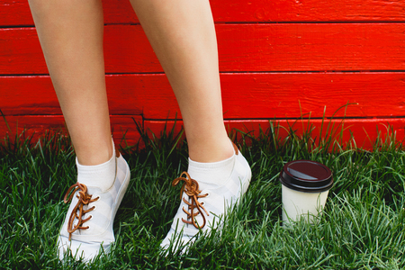 Woman's feet, in white socks and sneakers, standing on green grass near the cup of coffee, on red wooden background, close upの写真素材