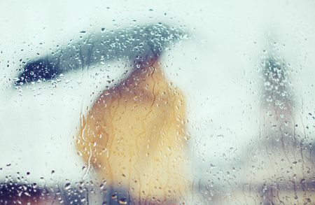 Man in yellow jacket with umbrella through glass with drops of water, on a background of city, rear viewの写真素材
