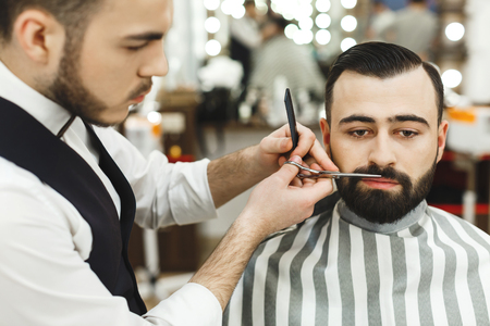 Barber with dark hair wearing white shirt making beard form with scissors for man with black hair at barber shop, mirror at background.の写真素材