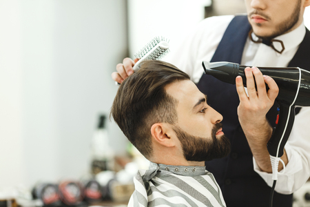 Man wearing white shirt doing a haircut with hair dryer and hair brush for man with black hair at barber shop, copy space, portrait.の写真素材