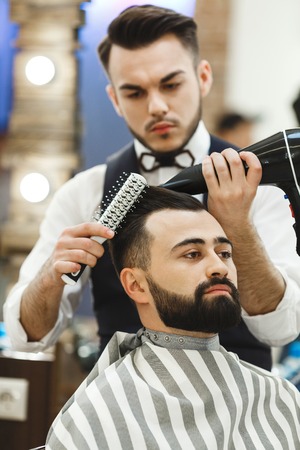 Barber with dark hair wearing white shirt doing a haircut with hair dryer and hair brush for man with black hair at barber shop, mirror at background.の写真素材