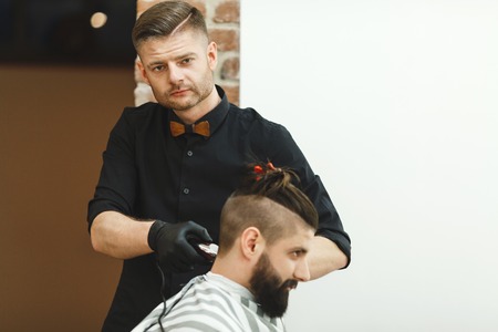 Handsome dark haired man wearing black shirt doing a haircut for man with long black hair and beard at barber shop, copy space, mock up.の写真素材