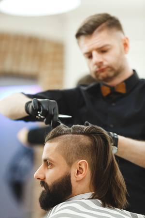 Attractive barber wearing black shirt, watch and black gloves doing a haircut for client with scissors at barber shop, portrait, copy space.の写真素材