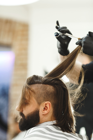 Man's hands in black gloves doing a haircut with scissors for man with dark long hair and beard at barber shop, close up portrait, copy space.の写真素材