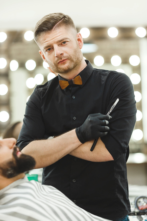 A portrait of handsome dark haired man wearing black shirt and gloves holding a razor at barber shop and looking at camera, mirrors at background.の写真素材