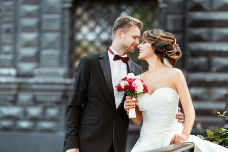 Wedding photo shooting. Bride and bridegroom standing with bouquet. Looking at each other and smiling. Woman wearing white dress and veil and man wearing suit. Outdoor, waist up, profileの写真素材