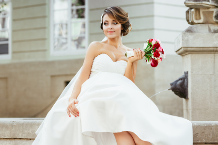 Wedding photo shooting. Bride sitting near monument in the city. Holding bouquet, looking aside. Wearing white dress, white shoes and veil. Outdoor, closeupの写真素材