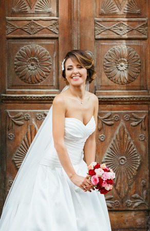Wedding photo shooting. Bride standing near wooden door and smiling. Wearing white dress and veil and holding bouquet. Outdoorの写真素材