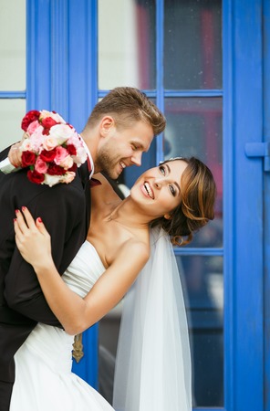 Wedding photo shooting. Bride and bridegroom standing near blue door and smiling. Embracing and leaning back, holding bouquet. Outdoor, closeup, waist upの写真素材