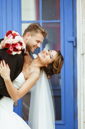 Wedding photo shooting. Bride and bridegroom standing near blue door and smiling. Embracing and leaning back, holding bouquet. Bride with closed eyes. Outdoor, closeup, waist upの写真素材