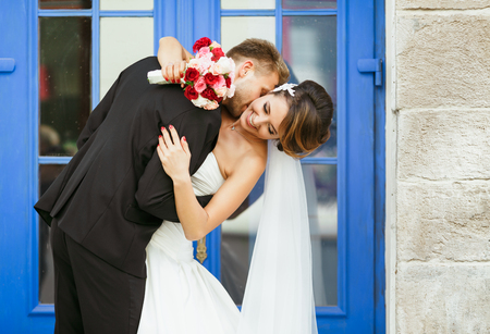 Wedding photo shooting. Bride and bridegroom near blue door. Embracing and leaning back, holding bouquet. Bridegroom kissing bride's neck and bride looking aside. Outdoorの写真素材