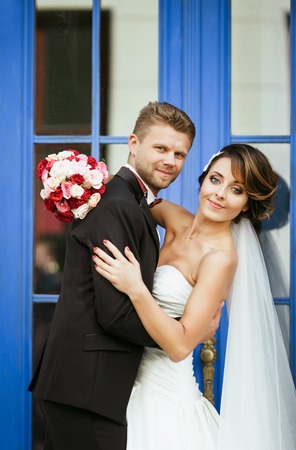 Wedding photo shooting. Bride and bridegroom near blue door. Embracing, holding bouquet. Looking at camera. Outdoorの写真素材