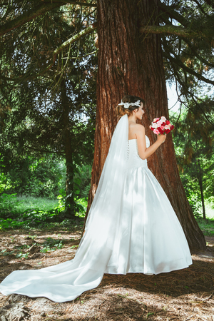 Wedding photo shooting. Bride standing in pine forest near tree with bouquet. Wearing white dress and long veil. Outdoor, full body, profileの写真素材