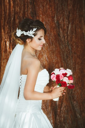 Wedding photo shooting. Bride standing in pine forest near tree with bouquet. Closed eyes. Wearing white dress and long veil. Outdoor, profileの写真素材
