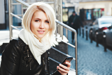 Beautiful blonde girl wearing black jacket and white scarf sitting outside and holding mobile phone, copy space.の写真素材