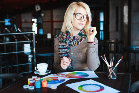 Cute blonde girl wearing gray blouse and scarf, sitting in cafe with cup of coffee and drawing, wearing glasses, paints and brushes on table.の写真素材