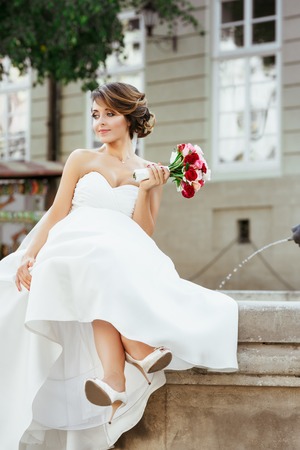 Beautiful bride sitting near monument and holding bouquetの写真素材