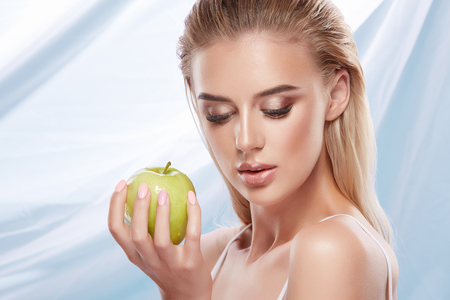 Beautiful girl with nude make up and blonde hair posing with green apple at bright backgroundの写真素材